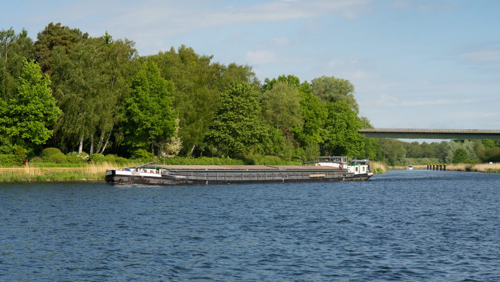 Das Binnenschiff Neptun passiert den Ziegelsee in Mölln. Im Hintergrund ist der weitere Verlauf des Kanals durch das Sportboot markiert; auf der Brücke überquert die B207 an dieser Stelle den Kanal. Das Foto entstand von der Kaikante des Möllner Stadthafens aus.