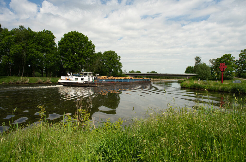 Das mit Stammholz für Lübeck voll beladene Frachtschiff Atlantis passiert bei Büchen den Parkplatz am Kanal samt Kunstinstallation. Hier verbringt manch einer bei gutem Wetter seine Mittagspause.
